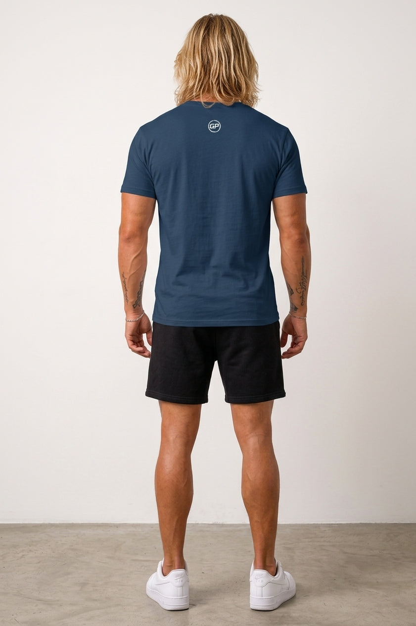 A man with long blonde hair is shown from behind wearing a navy blue t-shirt, with a white GP logo black shorts, and white sneakers against a plain off-white wall.