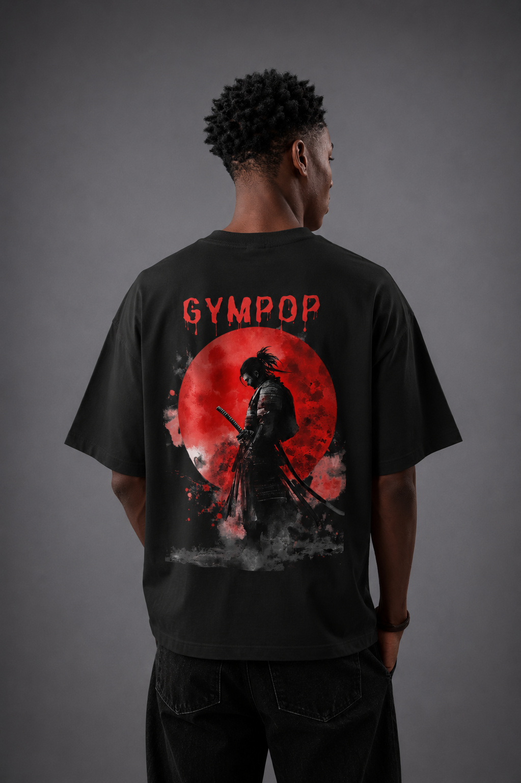 A man viewed from behind wearing a black t-shirt with a large graphic print on the back. The graphic features the text 'GYMPOP' in red above a samurai silhouette standing in front of a large red full moon, with dark smoke effects at the bottom.