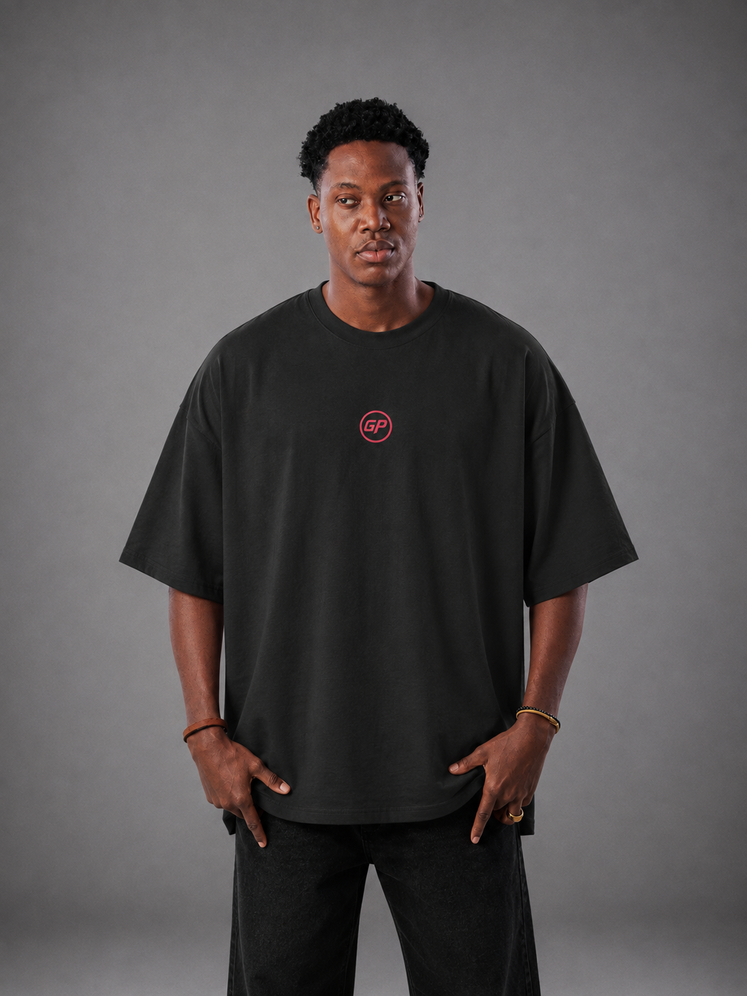 A man wearing an oversized black t-shirt with a small red circular GP logo on the left chest, paired with black pants, standing against a neutral grey studio background.