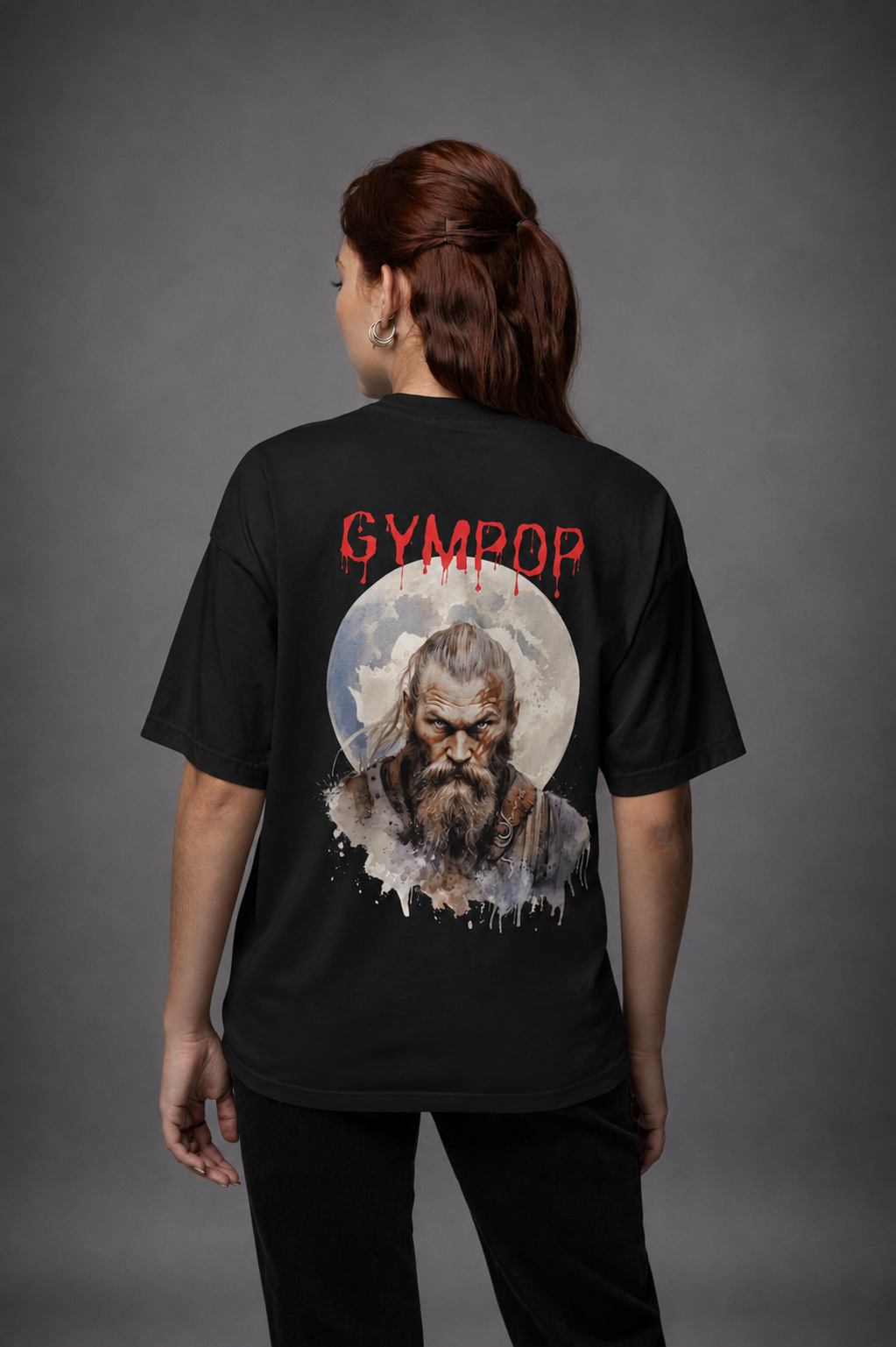 A woman viewed from behind wearing a black t-shirt with a large graphic print on the back. The graphic features the word 'GYMPOP' in red distressed lettering above a circular illustration of a bearded Viking with a ponytail in front of a full moon.