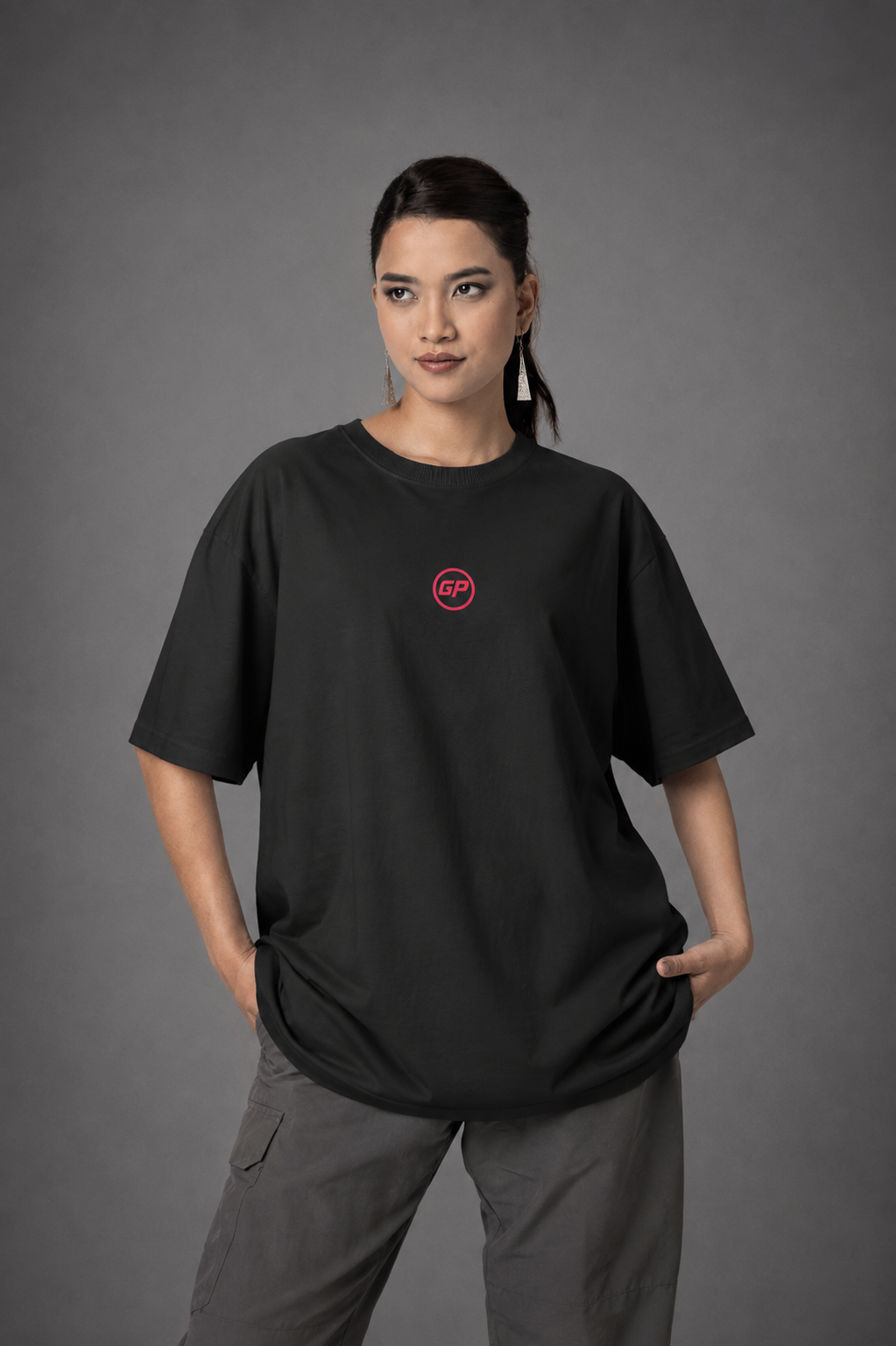 A woman modelling an oversized black t-shirt with a small red circular GP logo on the left chest, paired with grey cargo pants, against a dark grey studio background.