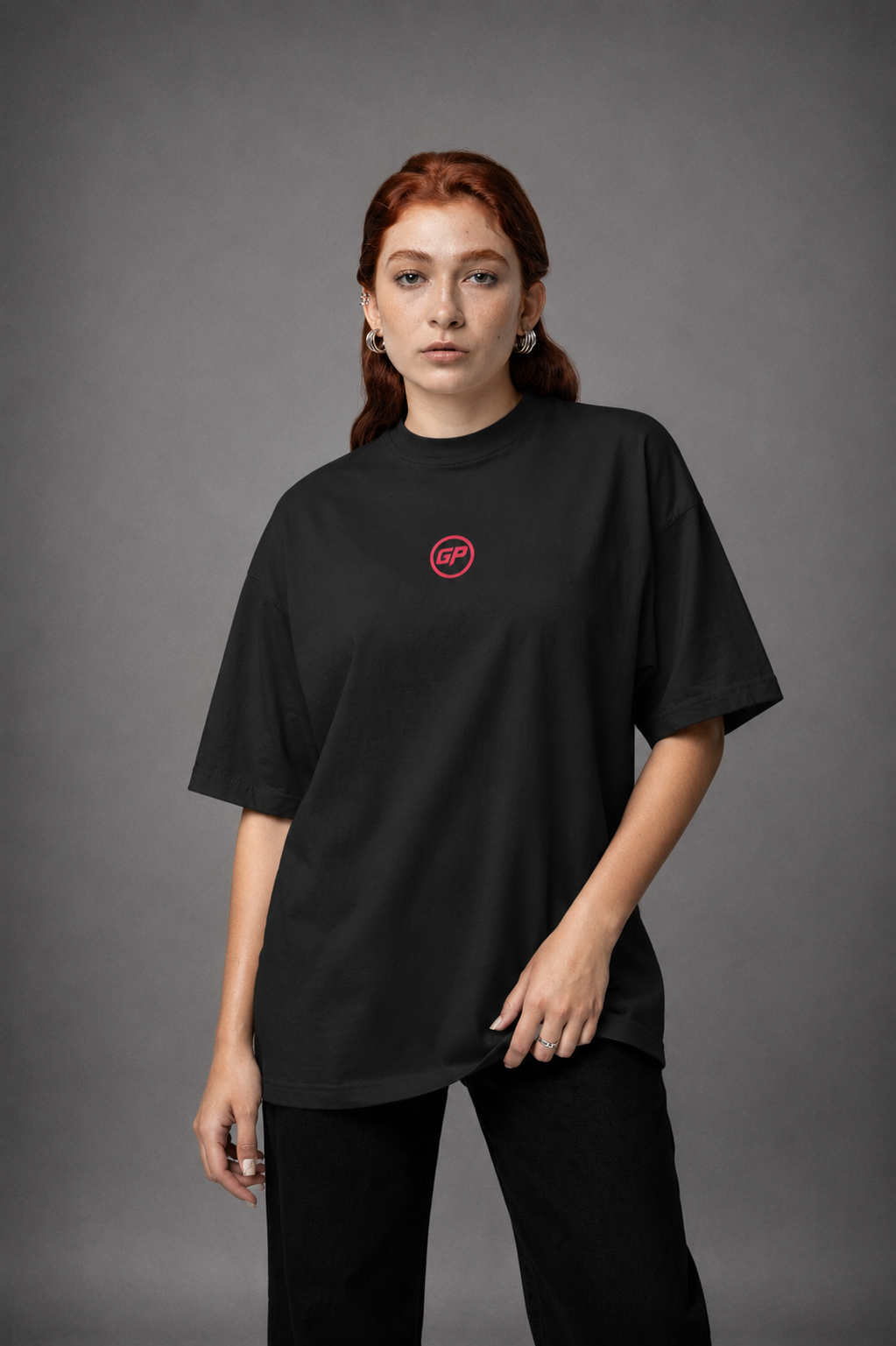 A model with red hair wearing an oversized black t-shirt with a small red circular GP  logo on the left chest, paired with black pants, against a dark grey studio background.