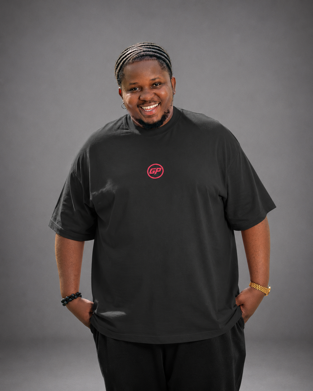 A man wearing a black short-sleeve t-shirt with a small red circular chest GP  logo, standing against a solid grey studio background.