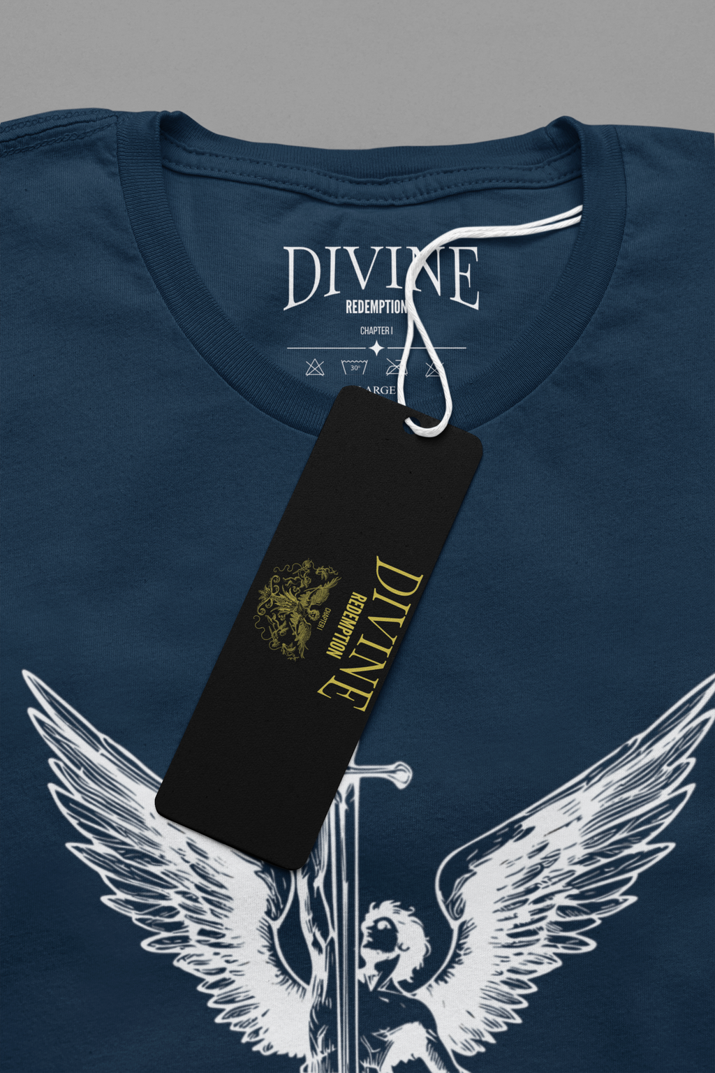 Close-up of a navy blue t-shirt with a white angel graphic holding a sword, featuring a black hangtag with gold 'DIVINE REDEMPTION' branding.