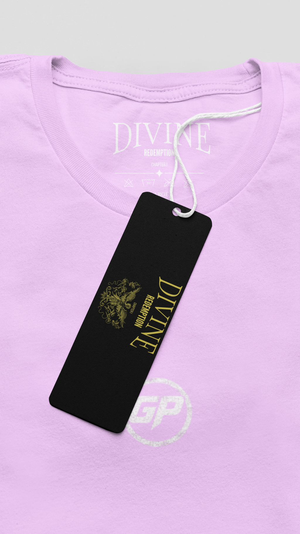 A close-up of a light Pink t-shirt with a black hangtag featuring gold branding. The brand 'DIVINE REDEMPTION' is printed in gold on the black tag, and the white 'DIVINE REDEMPTION' logo is also visible inside the collar.