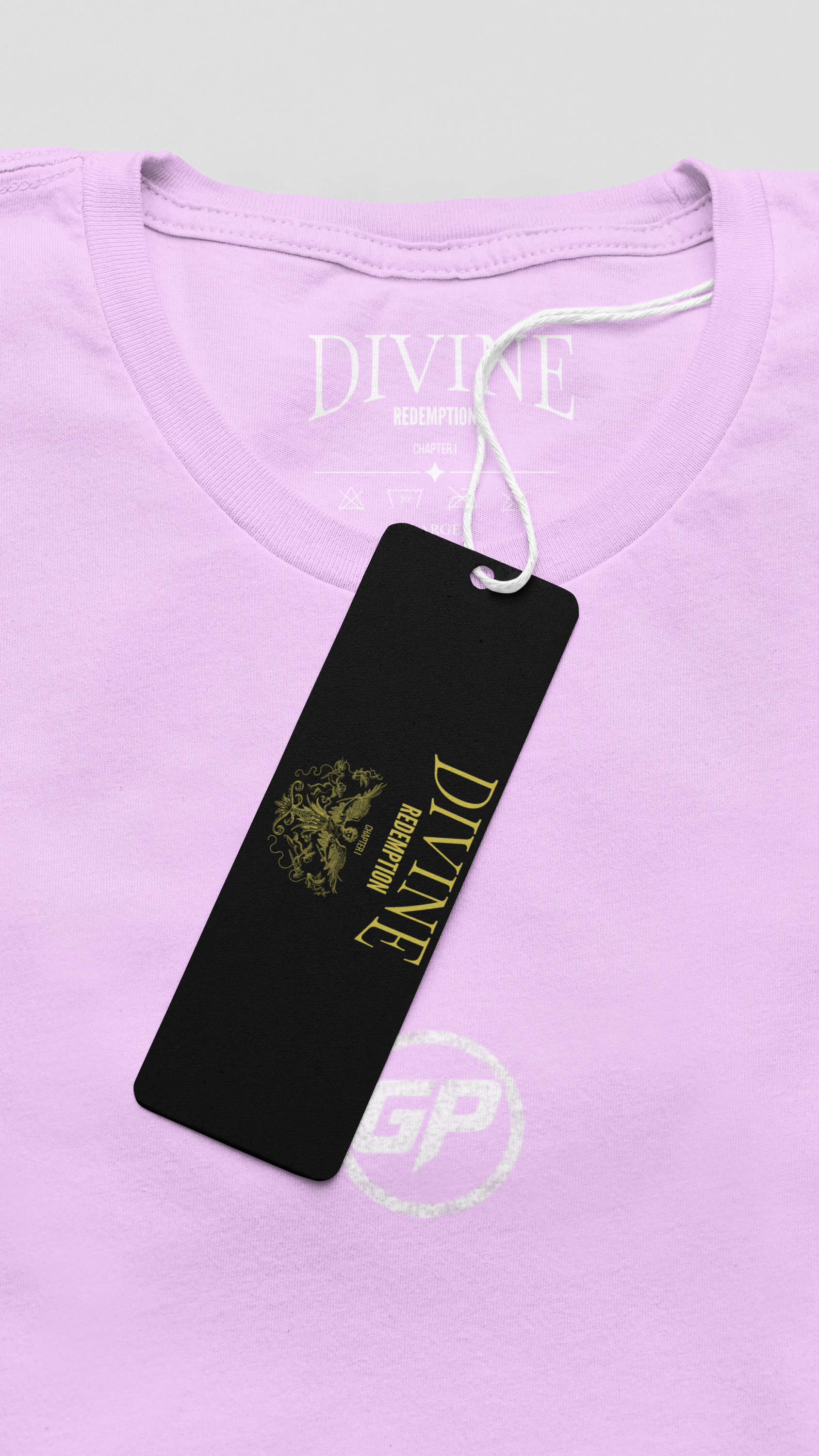 A close-up of a light Pink t-shirt with a black hangtag featuring gold branding. The brand 'DIVINE REDEMPTION' is printed in gold on the black tag, and the white 'DIVINE REDEMPTION' logo is also visible inside the collar.