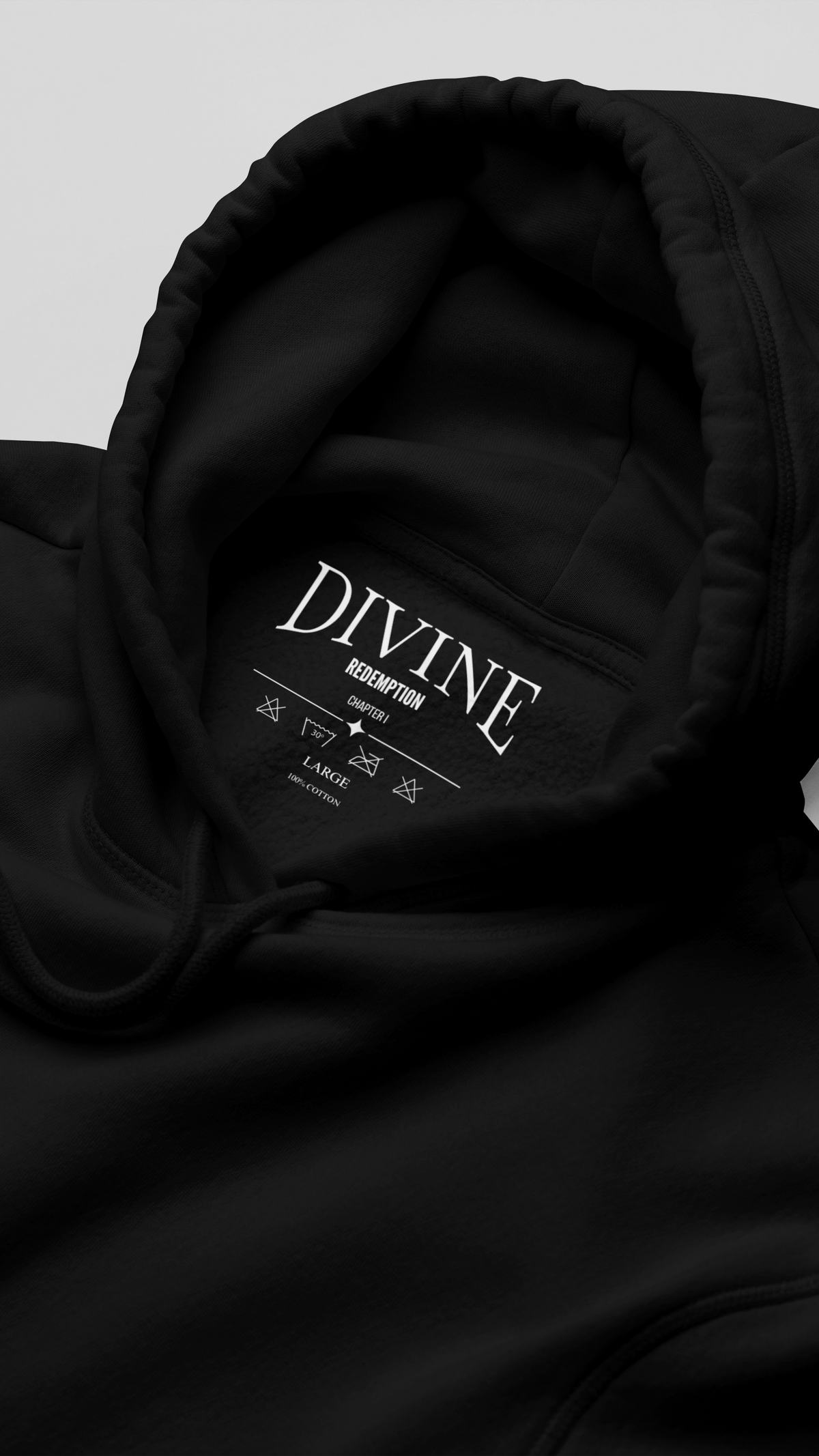 Close-up of a black hoodie hood with white printed text reading 'DIVINE REDEMPTION' and care instructions inside the collar, photographed on a light grey background.